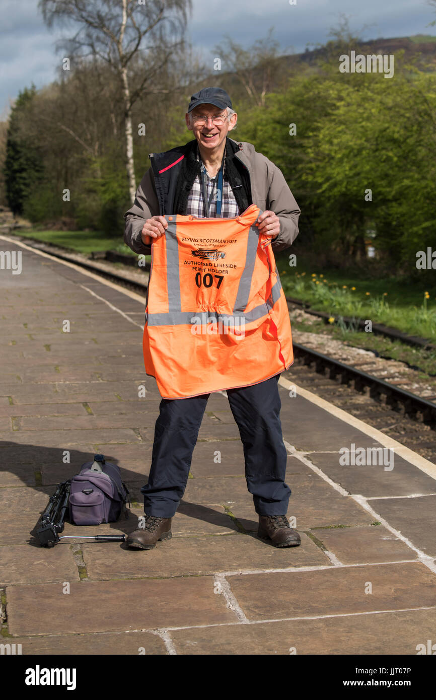 Smiling man standing on station platform, posing & holding hi-vis jacket before arrival of Flying Scotsman - Keighley & Worth Valley Railway, GB, UK. Stock Photo