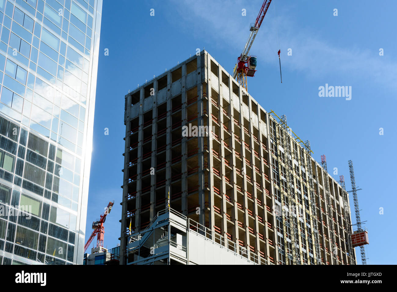 Low angle view of a concrete building under construction next to a glass building with two tower cranes against blue sky. Stock Photo