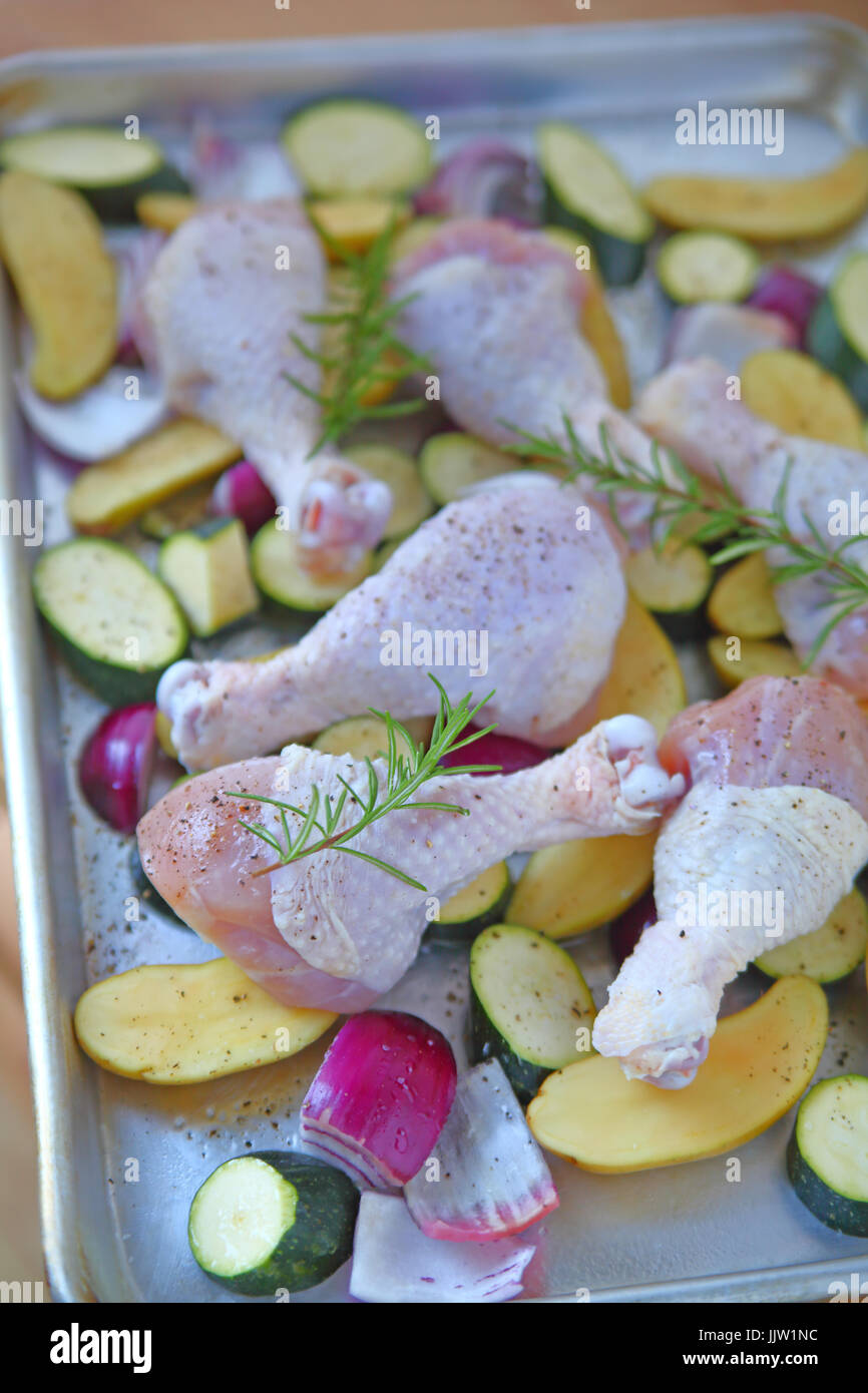 Baking sheet with chicken legs, zucchini, rosemary, onions, and potatoes before going in the oven Stock Photo