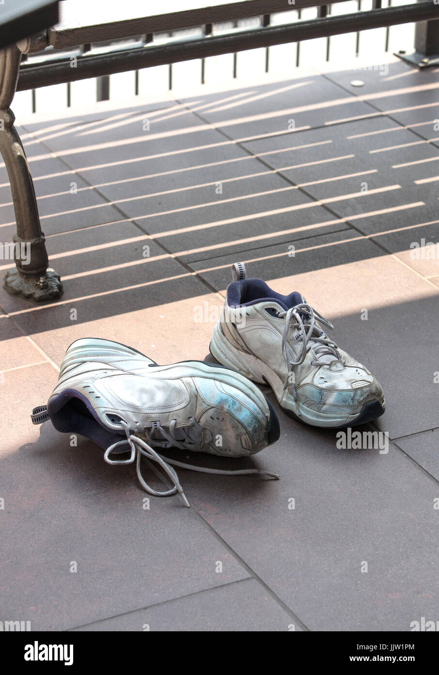 A pair of old and used running shoes laying on a tiled concrete street floor next to a bench Stock Photo