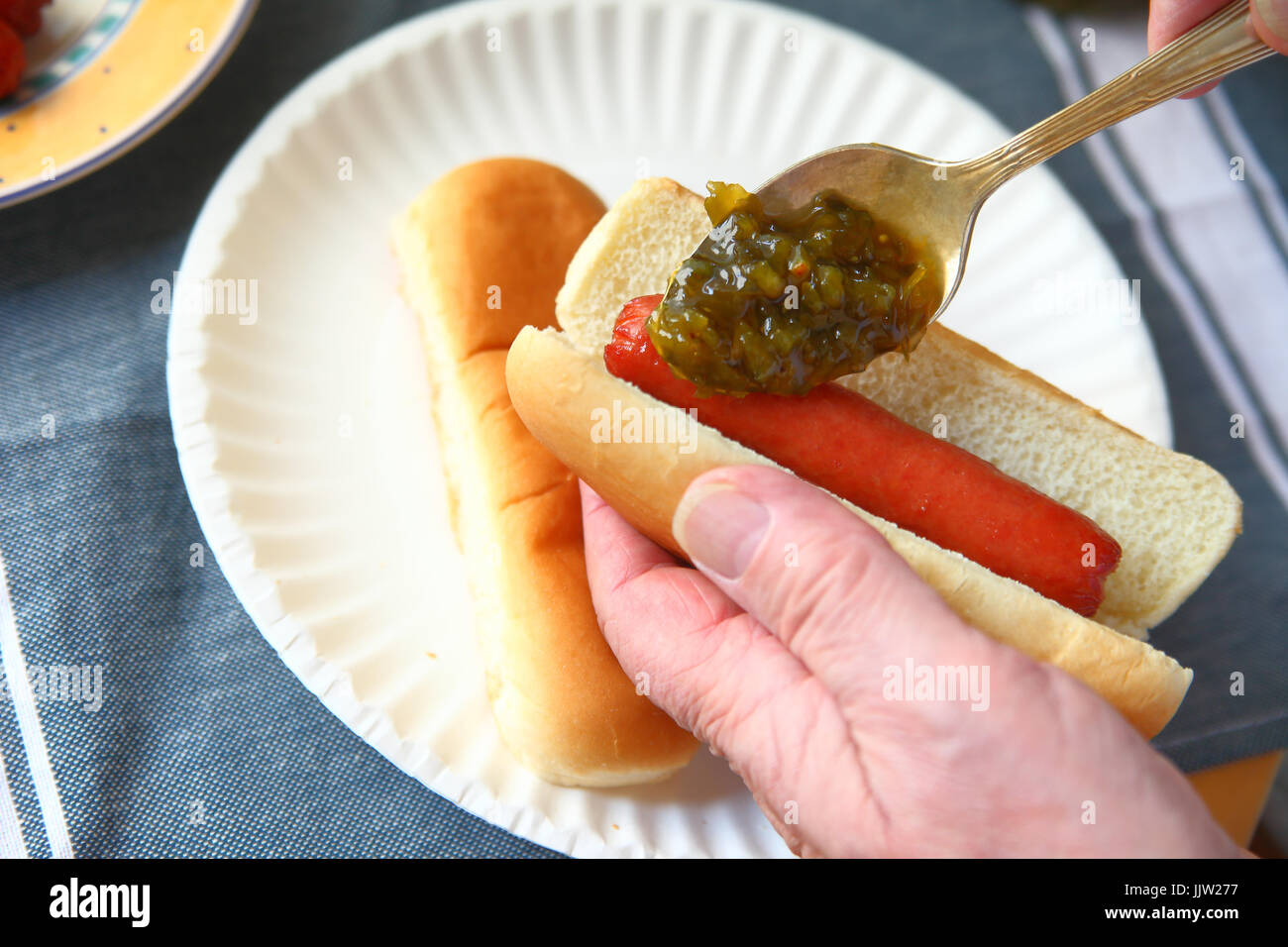 A man adds sweet pickle relish to a hot dog over a paper plate with