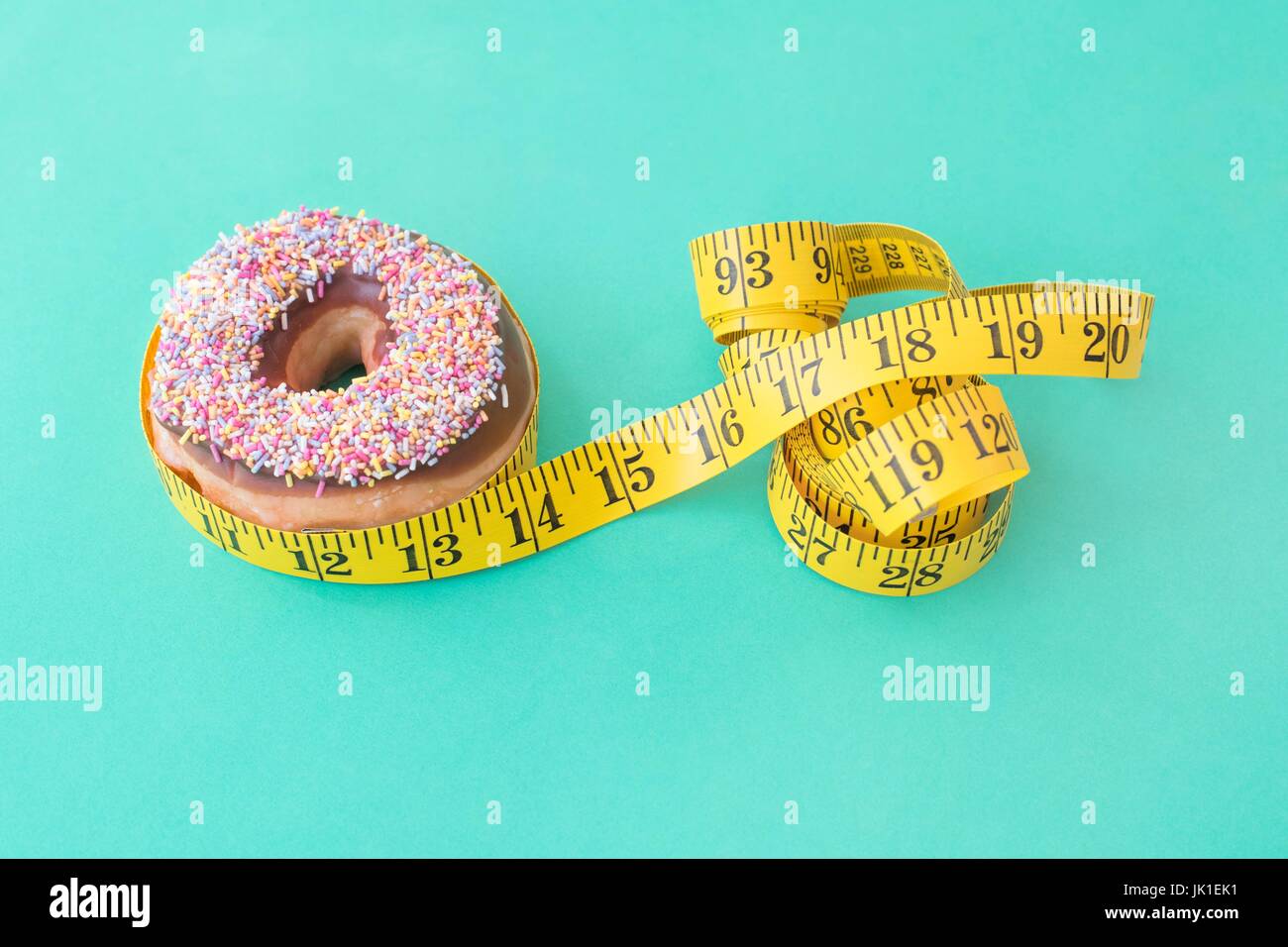 Doughnut with sprinkles and yellow tape measure against a green background. Conceptual image of controlling calorie intake. Stock Photo
