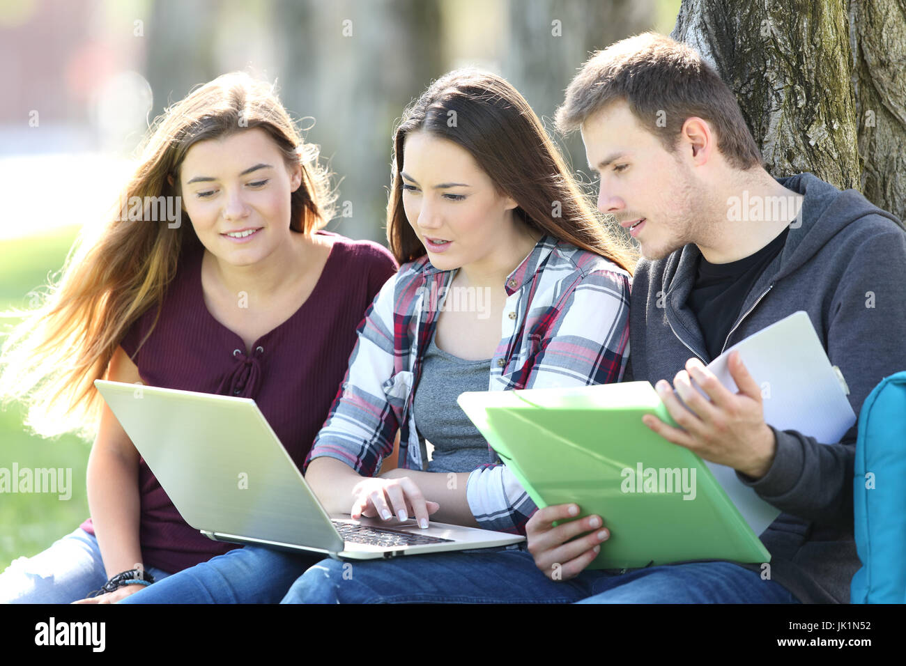Three attentive students learning on line together with a laptop and notebook sitting on the grass in a park Stock Photo