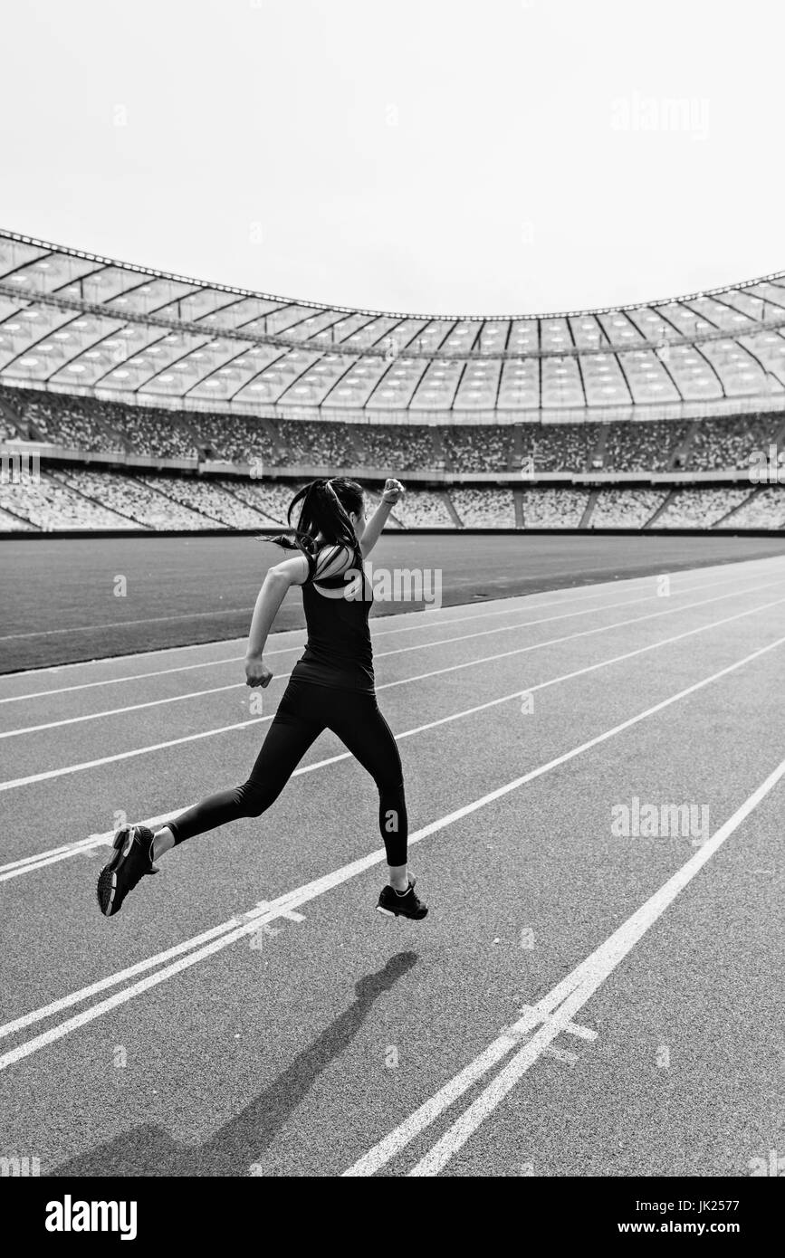 Back view of young fitness woman in sportswear sprinting on running track stadium, black and white photo Stock Photo