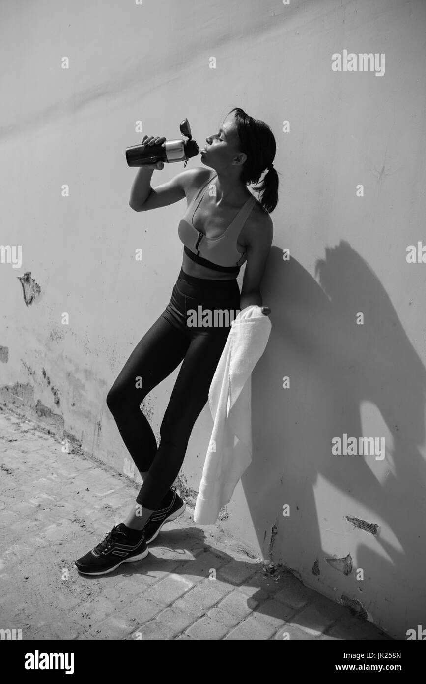 Tired young sportswoman leaning on wall at stadium and drinking water from sports bottle, black and white photo Stock Photo