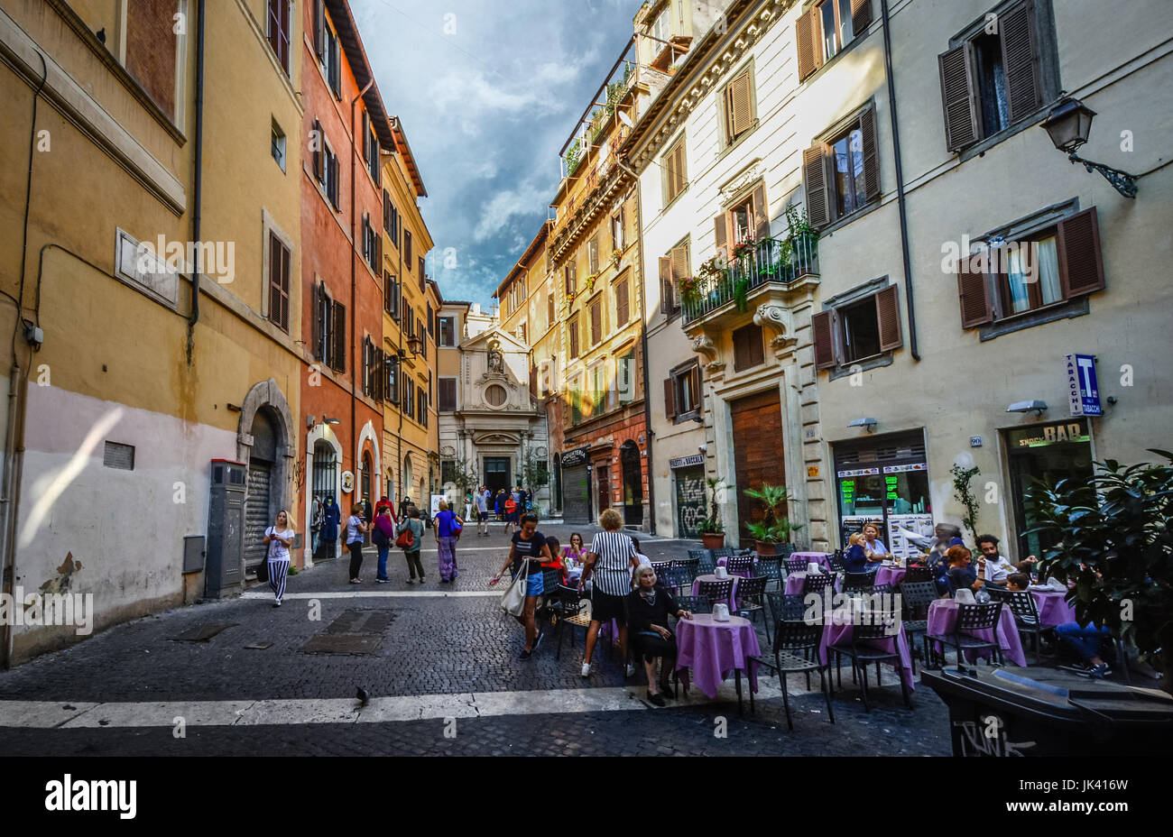 Small out of the way piazza off the beaten path in Rome Italy with an old woman sitting alone at a cafe while younger italians go about their day Stock Photo