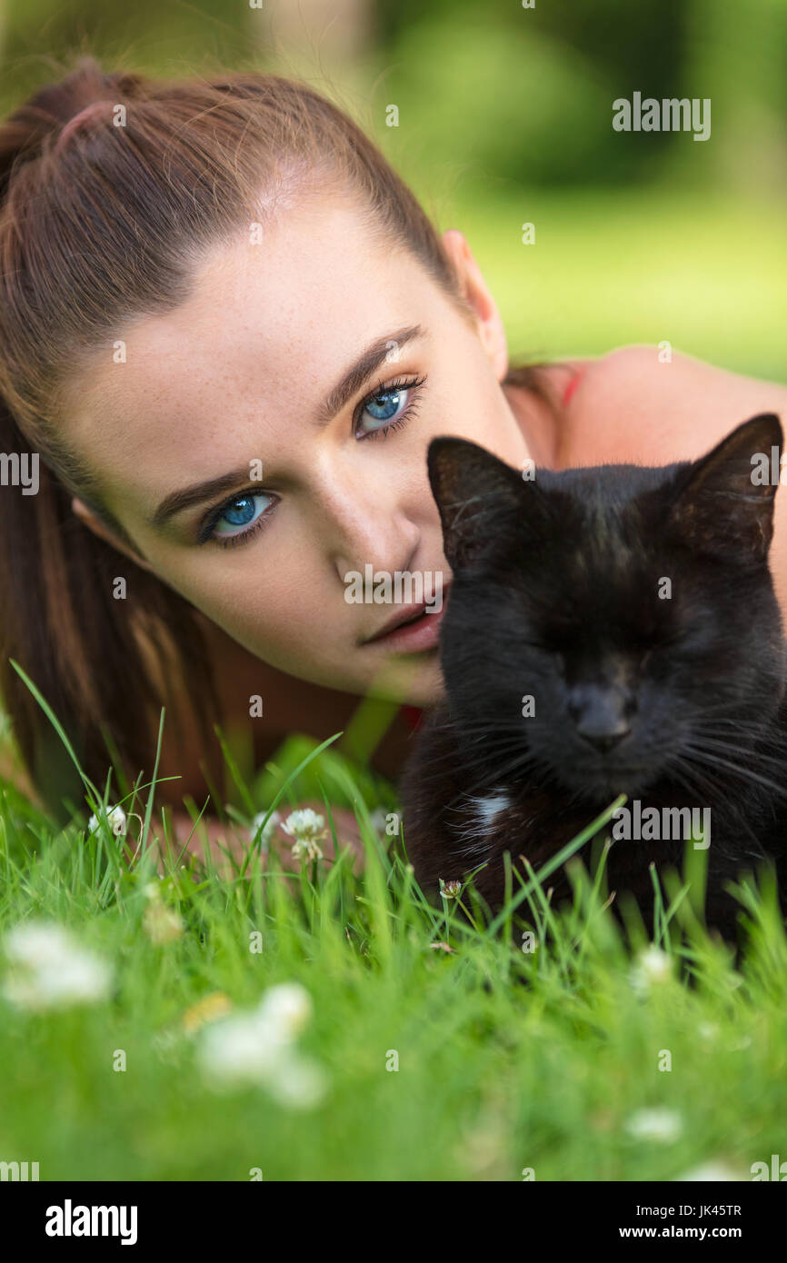 Beautiful female teenager girl young woman with blue eyes, laying down outside on grass with a black cat Stock Photo