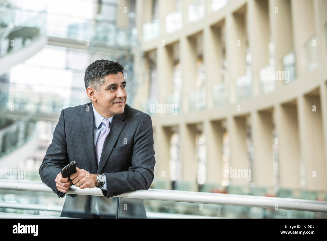 Mixed Race businessman leaning on railing in lobby holding cell phone Stock Photo