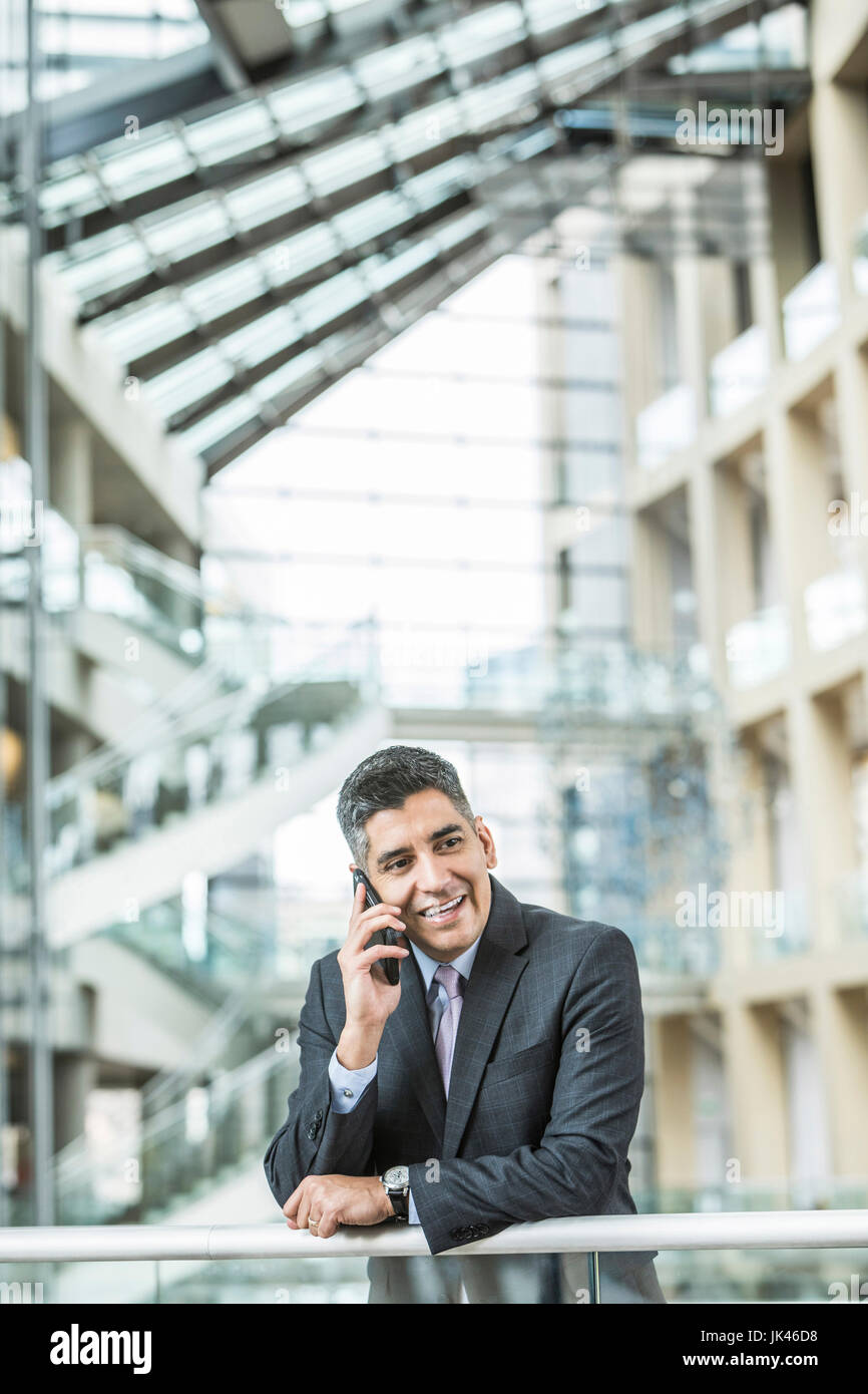 Mixed Race businessman leaning on railing in lobby talking on cell phone Stock Photo