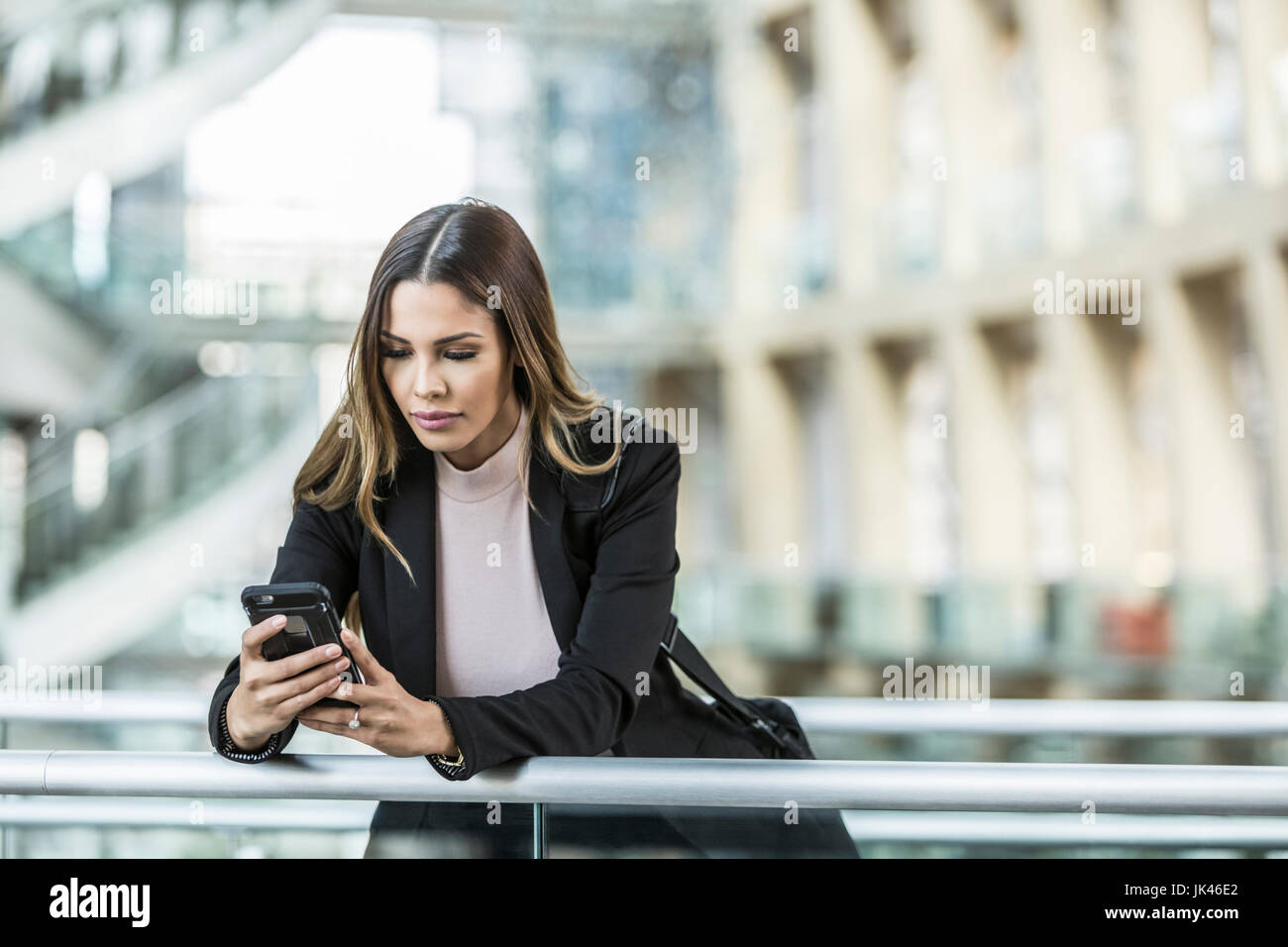Mixed Race businesswoman texting on cell phone leaning on railing in lobby Stock Photo