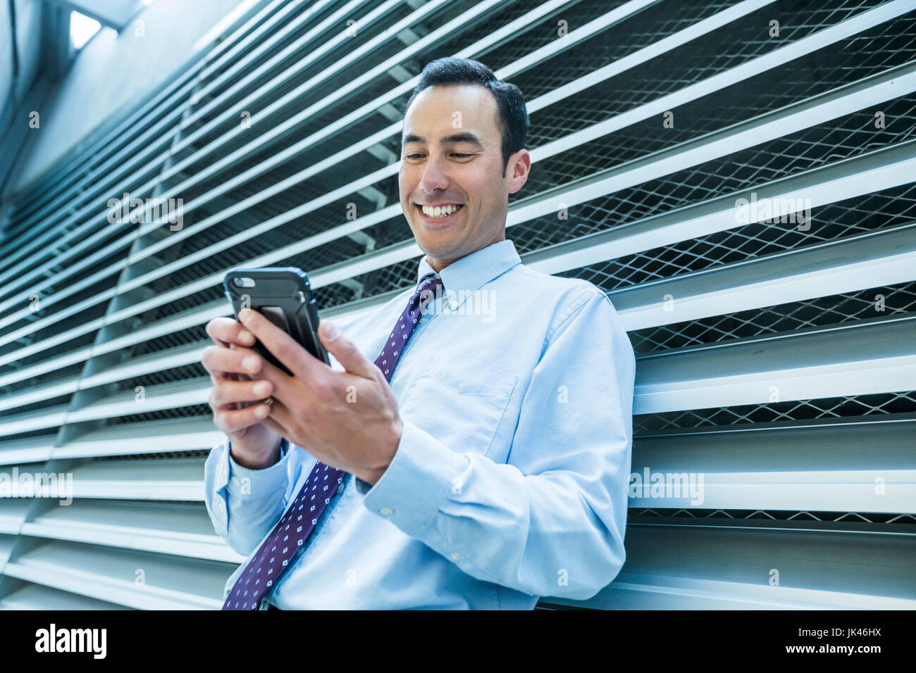 Smiling Mixed Race businessman leaning on wall texting on cell phone Stock Photo