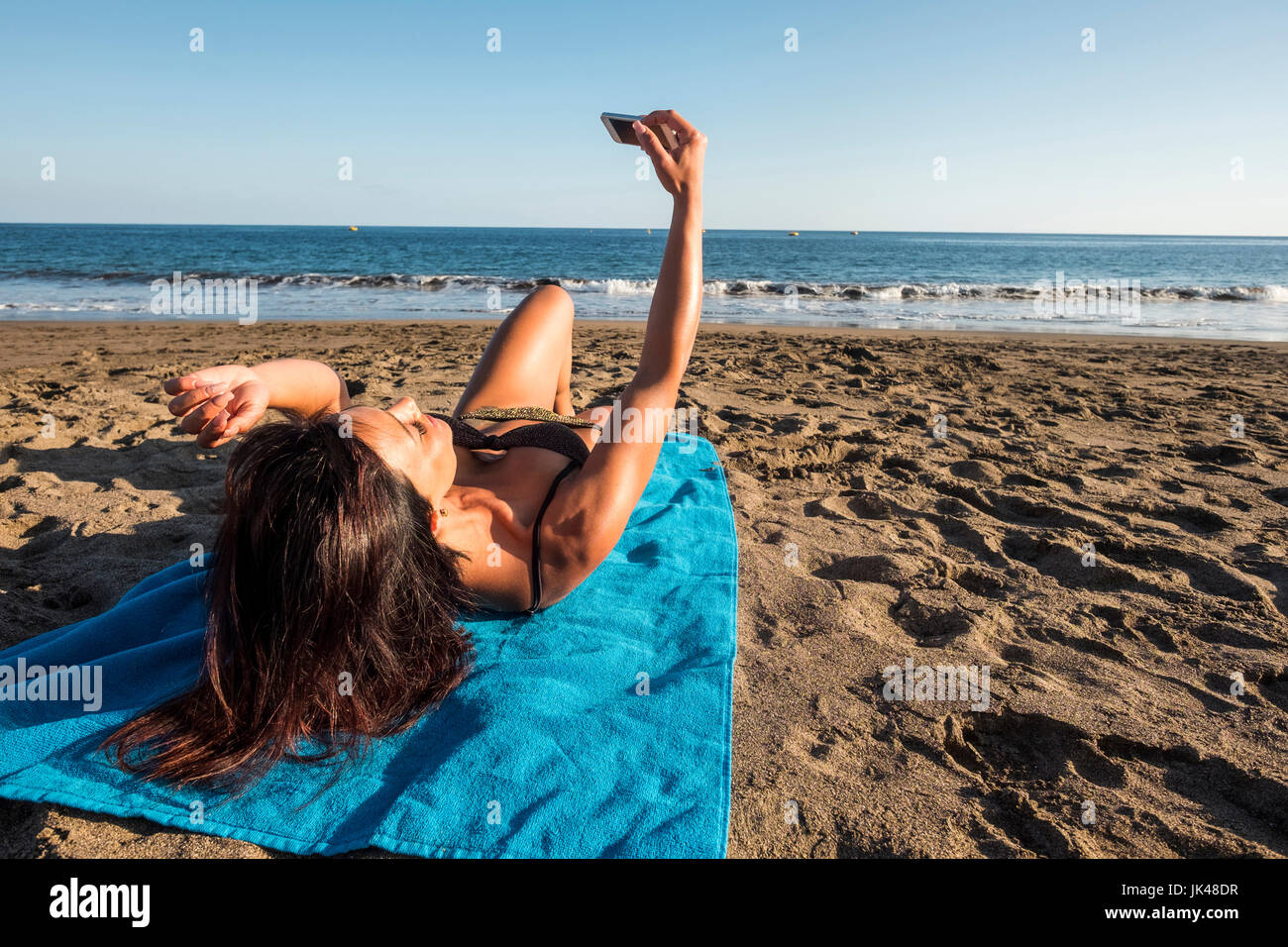 Caucasian woman laying on blanket on beach posing for cell phone selfie Stock Photo