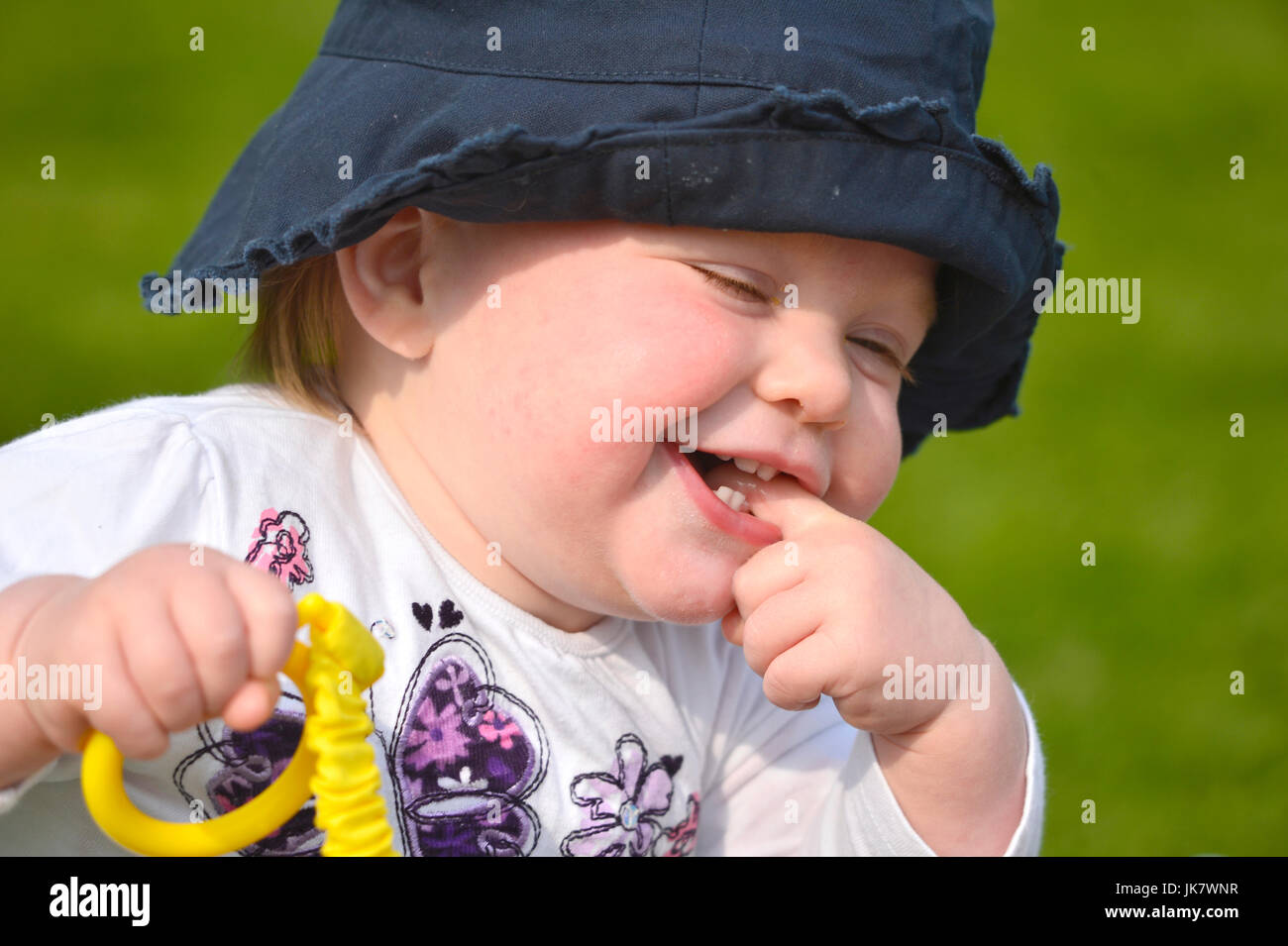 baby girl with finger in mouth wearing blue hat holding rattle Stock Photo