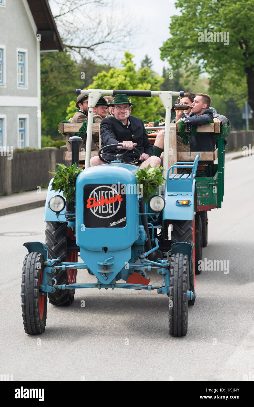 Young men and women in Bavarian costumes sitting on a trailer pulled by an old tractor arriving at a traditional may celebration Stock Photo