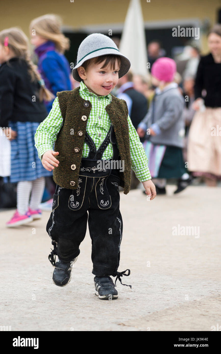 Child in traditional Bavarian dress with leather trousers, cardigan and fur hat sticking his tongue out walking on the dance floor around a maypole Stock Photo