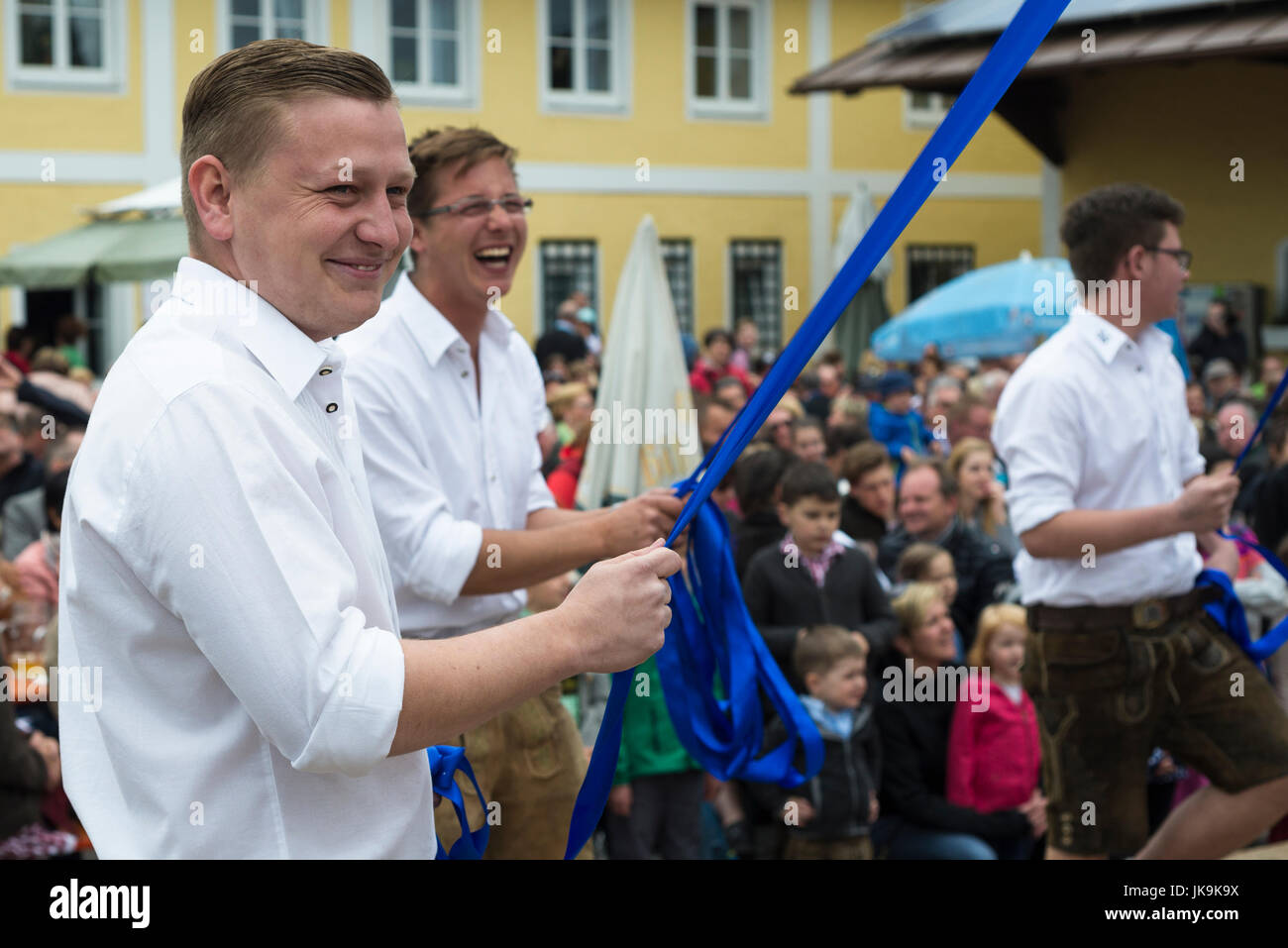 Young Bavarian men in white shirt and leather trousers holding blue ribbon while performing traditional folk dance Bandltanz around the maypole Stock Photo