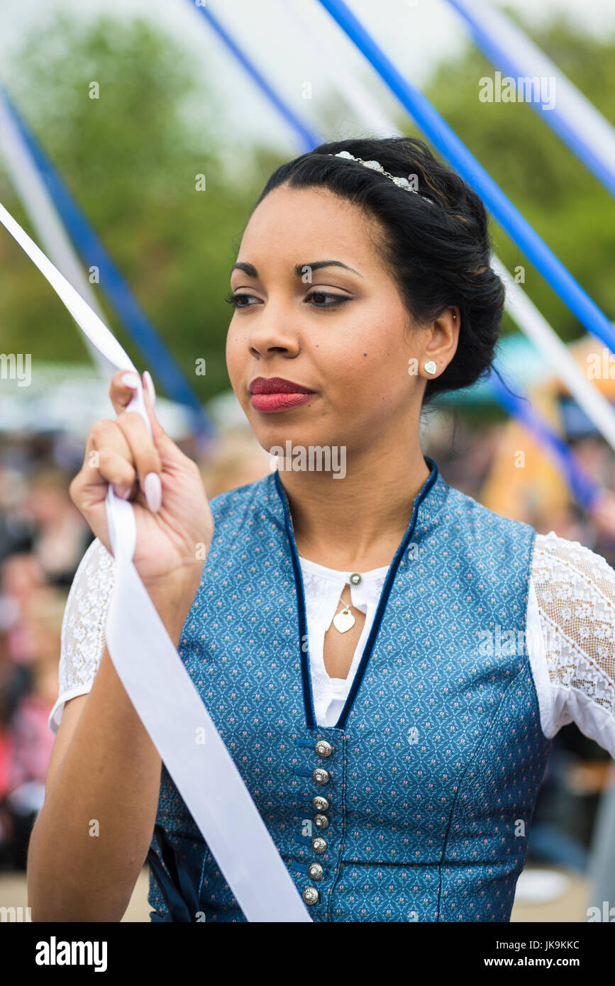 Young woman in traditional dirndl dress holding white ribbon while performing traditional folk dance Bandltanz around the maypole Stock Photo