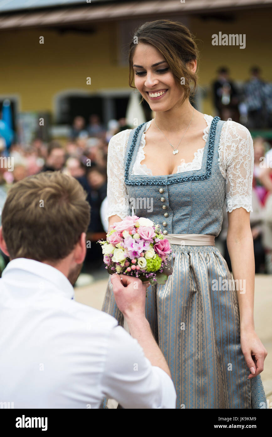 Portrait of a young man kneeling in front of a woman with a bouquet of flowers performing a traditional Bavarian folk dance Bandltanz around a maypole Stock Photo