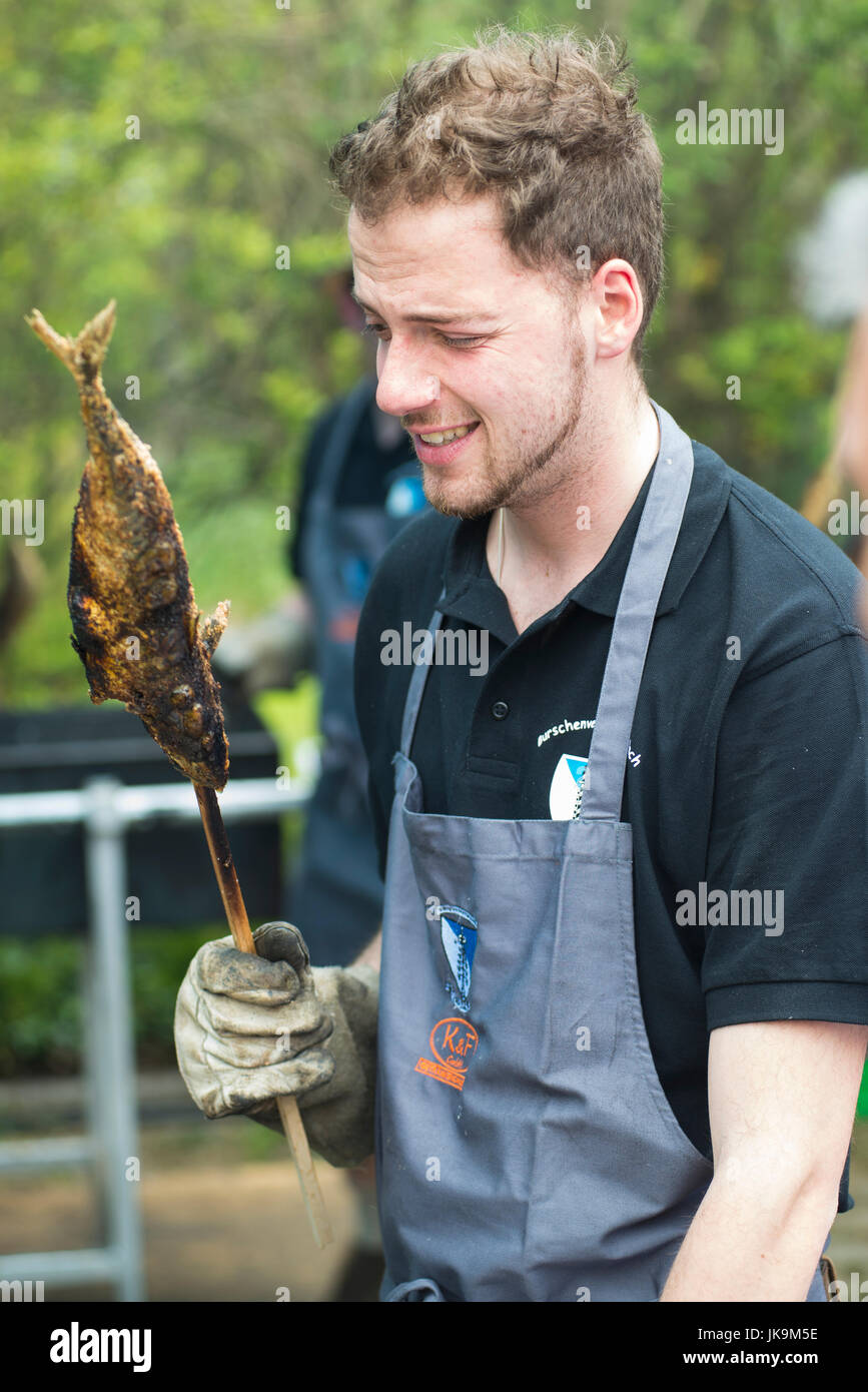 Young man frowning and holding a fish on a stick like trout,char or whitefisch grilled above fervid coal at a traditional barbecue food in Bavaria Stock Photo