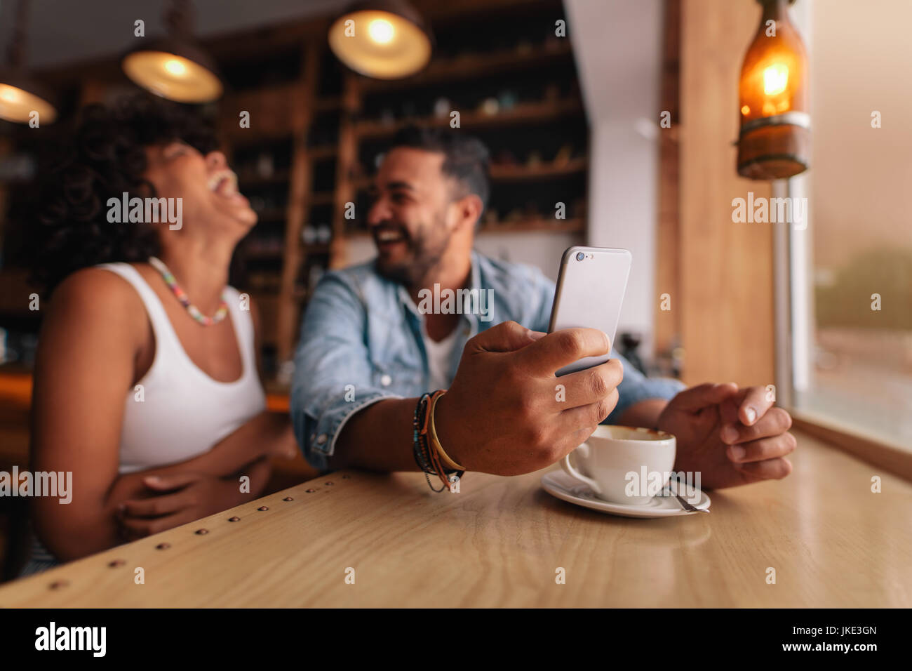 Young couple enjoying at coffee shop. Man holding mobile phone with laughing woman sitting by. Stock Photo