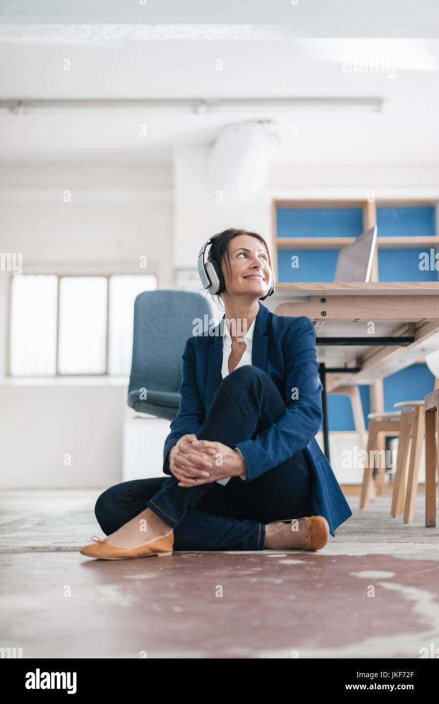 Smiling businesswoman sitting on the floor in a loft listening music ...