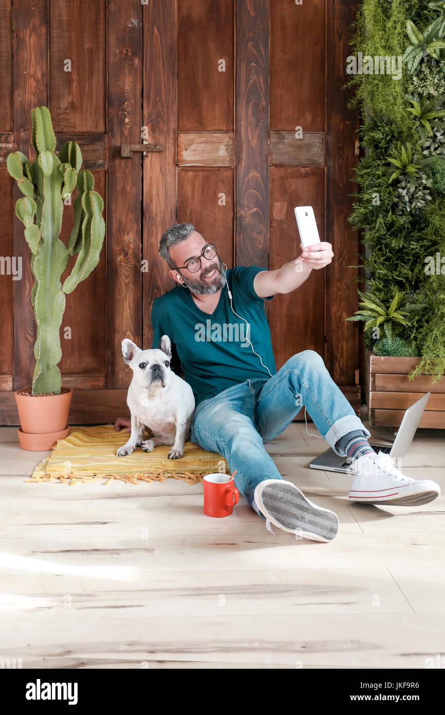 Bearded man sitting with his dog on the floor at home taking selfie with smartphone Stock Photo