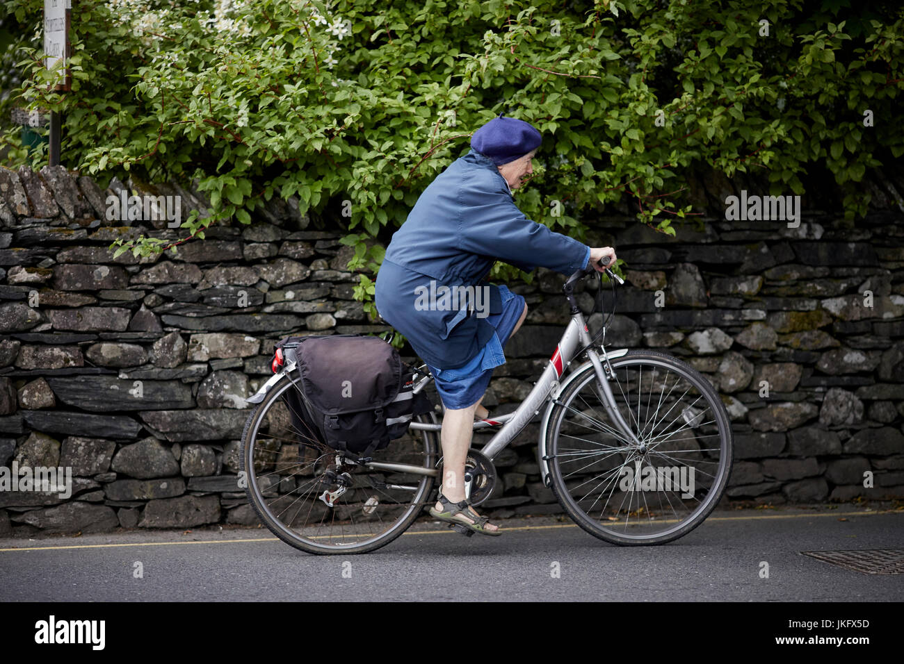 A old lady cycles through Grasser village in Cumbria’s Lake District National Park. Stock Photo