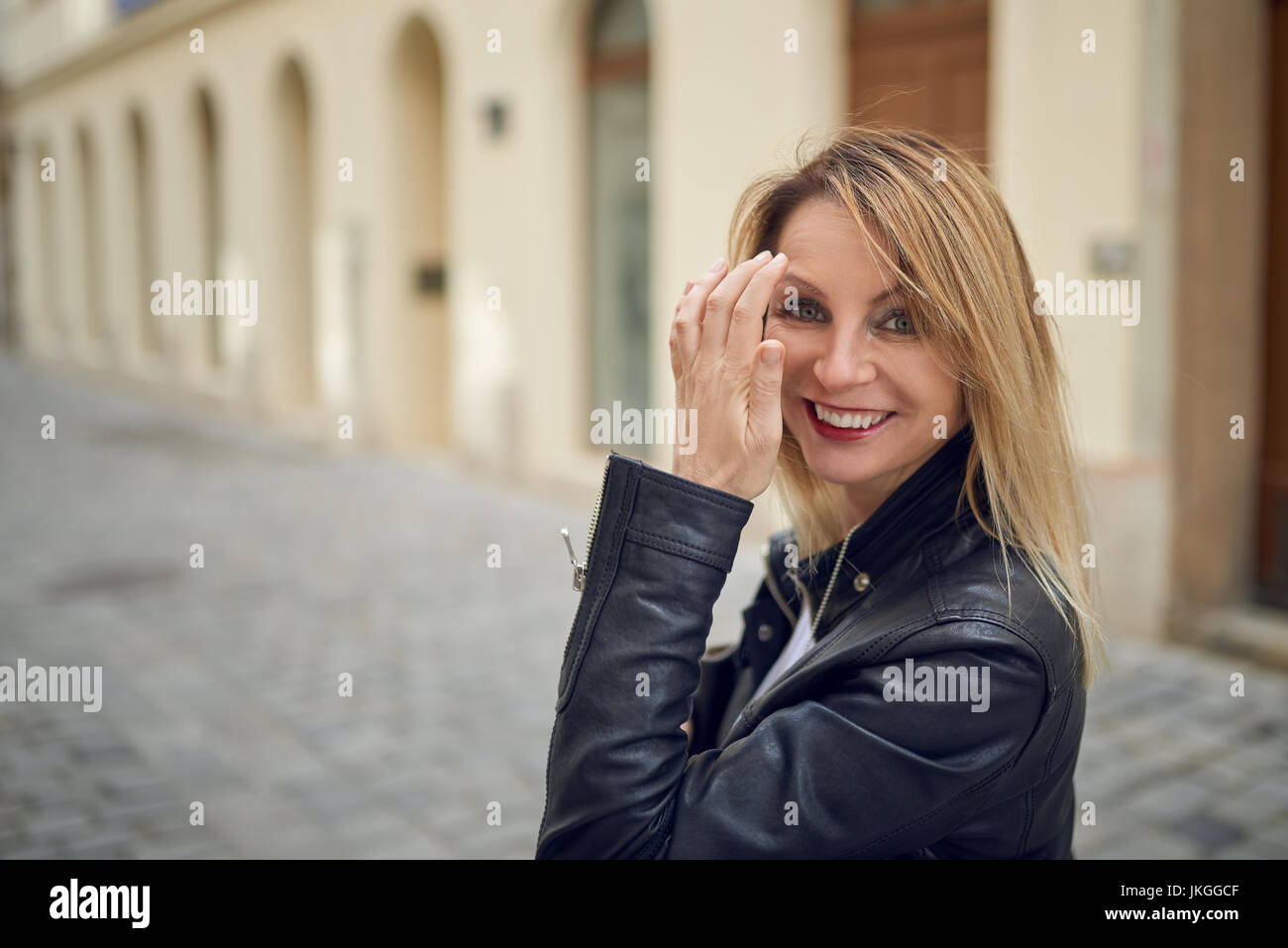 Attractive blond woman standing outdoors in a courtyard flicking her long blond hair off her face with her hand as she smiles at the camera Stock Photo