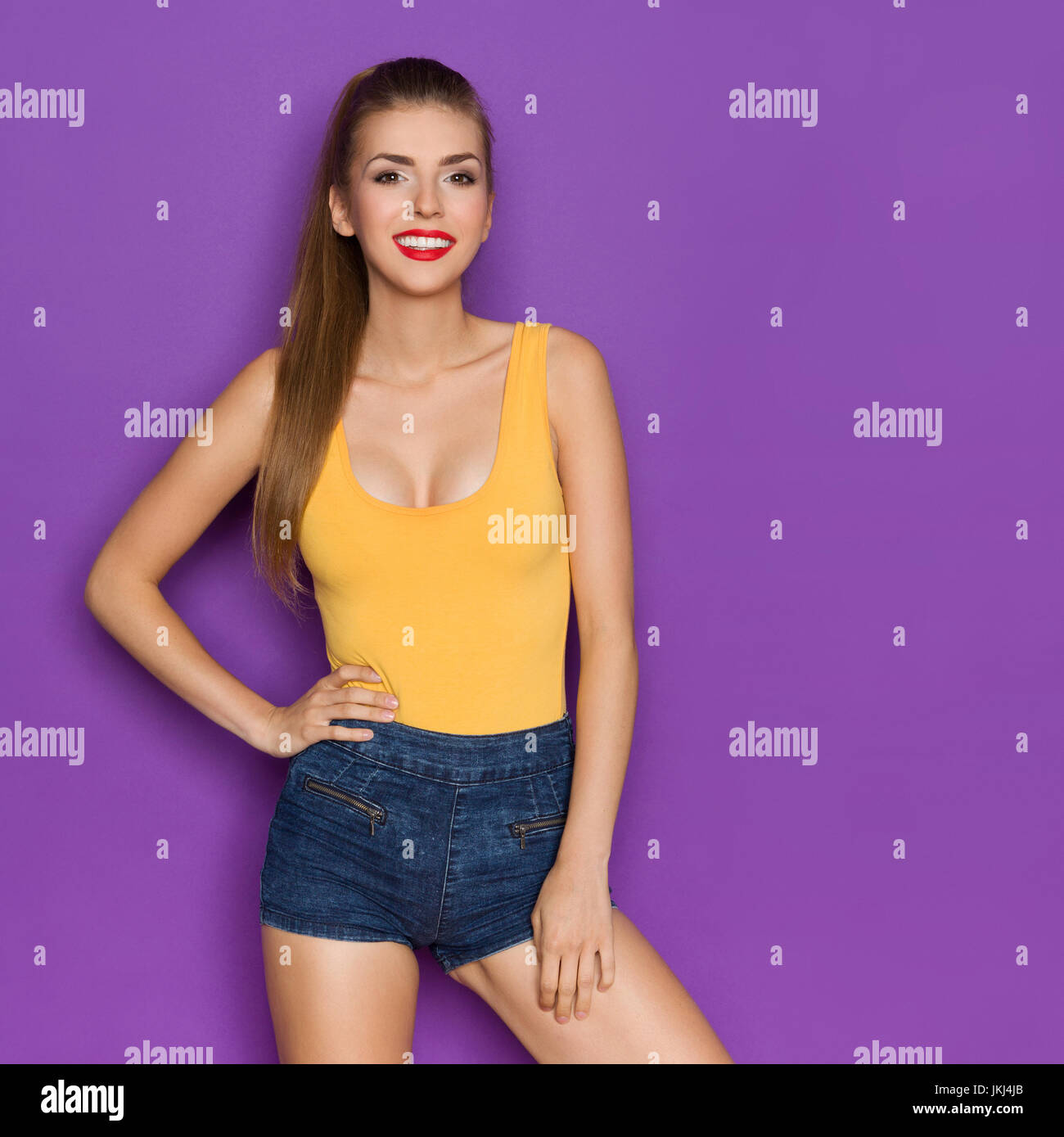 Smiling young woman in orange shirt and jeans shorts posing with hand on  hip. Three quarter length studio shot on purple background Stock Photo -  Alamy