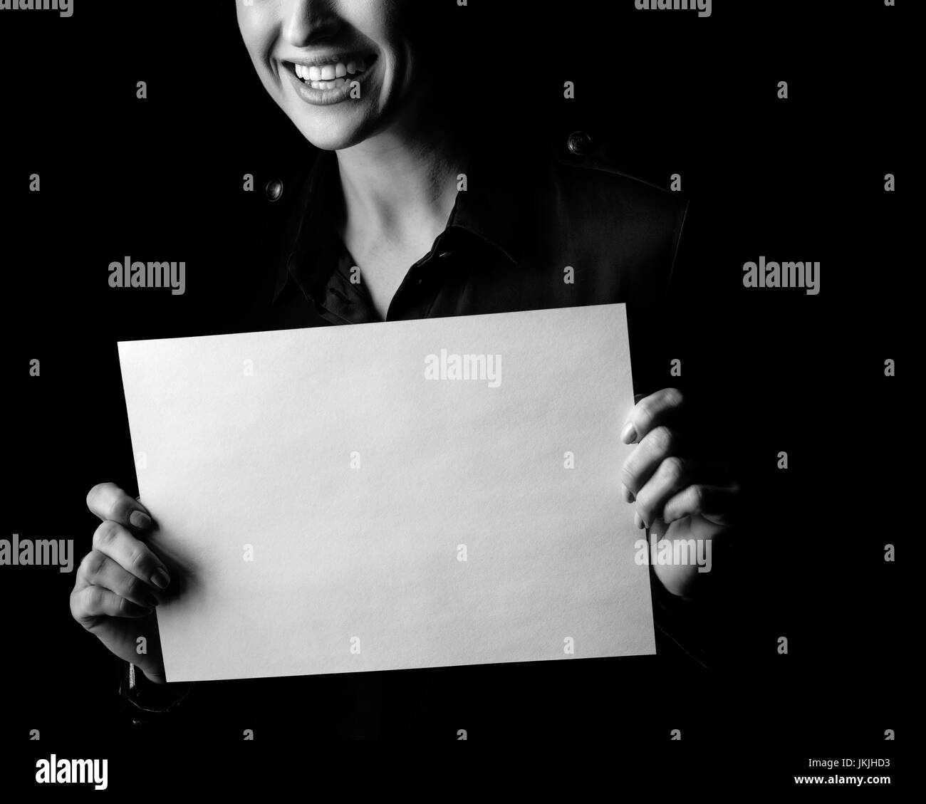 Сoming out into the light. Closeup on smiling woman in the dark dress isolated on black background showing blank paper sheet Stock Photo