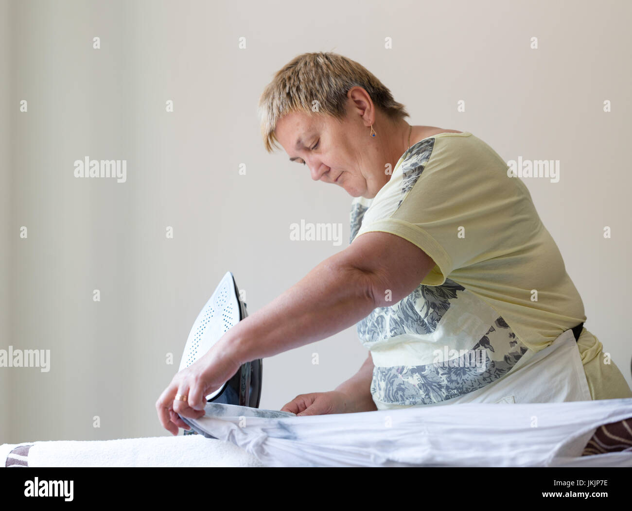 Senior woman ironing washed cloths in light room. Horizontal orientantion with selective focus on working hands with black iron. Direct front view Stock Photo