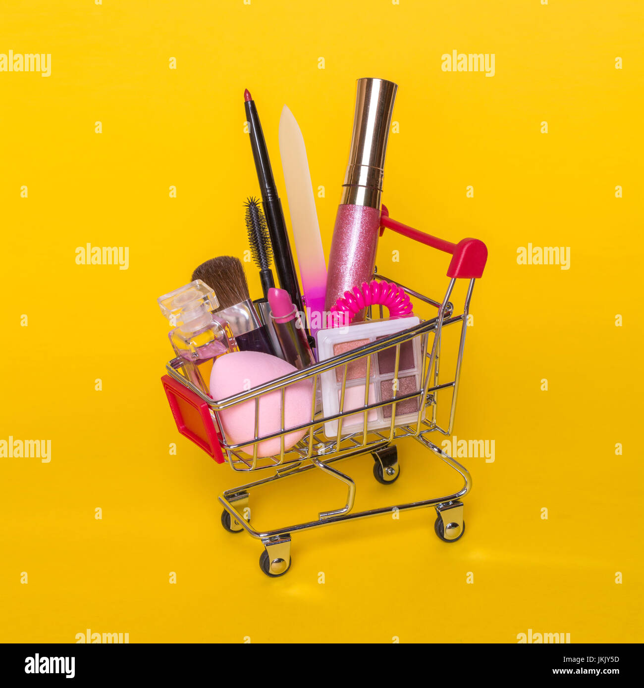 Creative concept with shopping trolley with makeup on a yellow background. Perfume, sponge, brush, mascara, pencil, nail file, eye shadow, lip gloss i Stock Photo