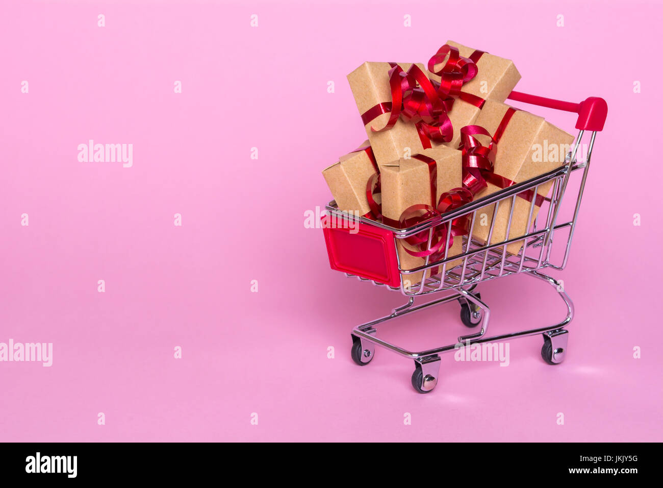 Creative concept with shopping trolley with gifts on a pink background. Gifts wrapped in kraft paper with a red ribbon and bow. Stock Photo