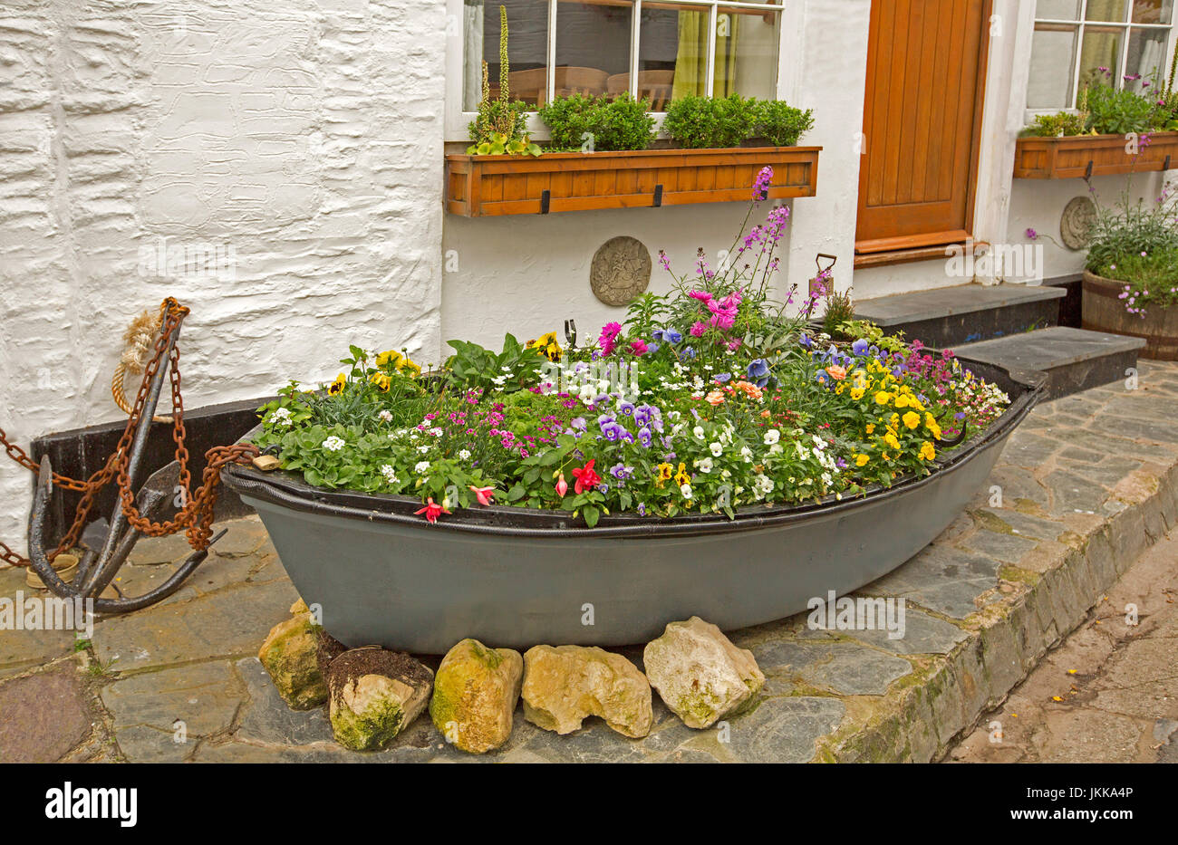 Colourful mass of flowering annuals, including pansies and petunias, growing in a boat - an old dinghy, beside the door and white wall of house Stock Photo