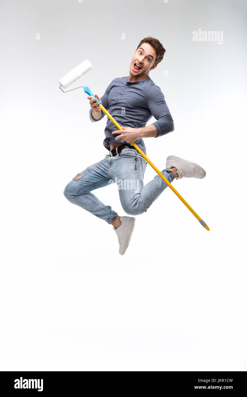 handsome man holding paint roller and jumping isolated on white