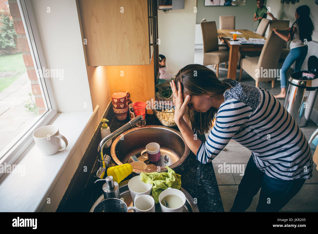 Stressed mum at home. She has her head in her hands at a messy kitchen sink and her children are running round in the background. Stock Photo