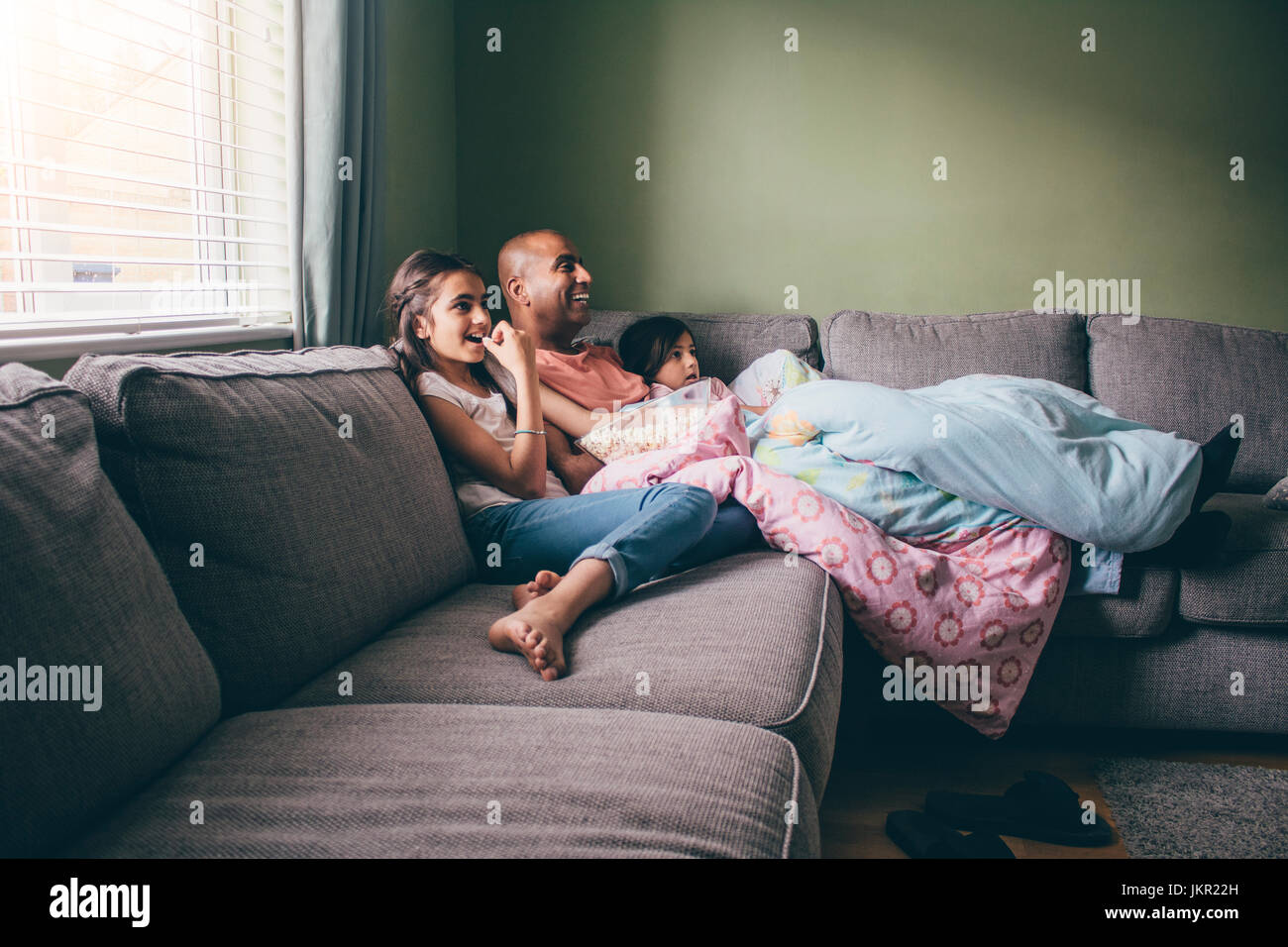 Father curled up on the sofa with his two daughters. They are watching a movie with popcorn. Stock Photo