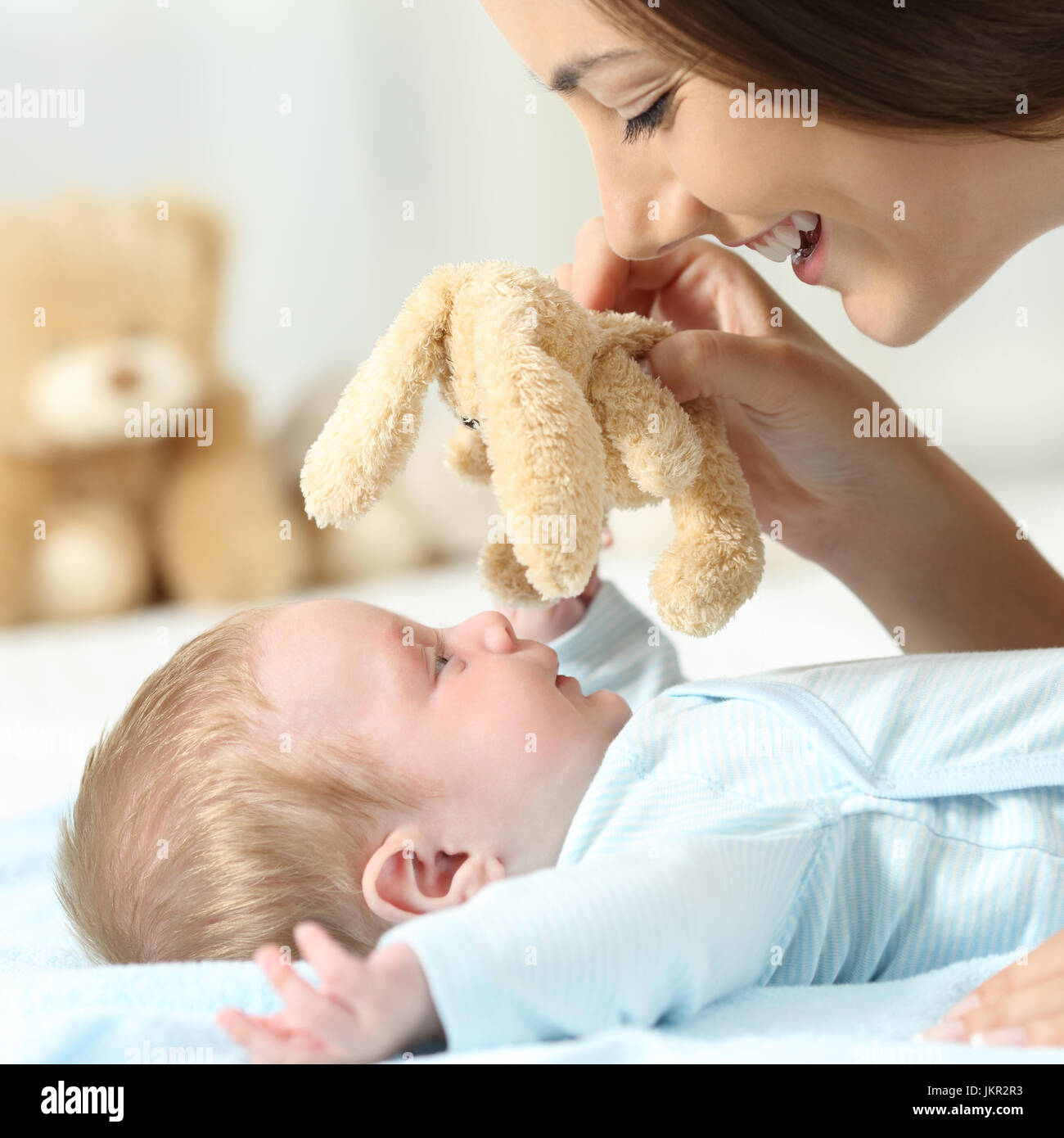 Side view close up portrait of a mother playing with her baby holding a teddy on a bed Stock Photo