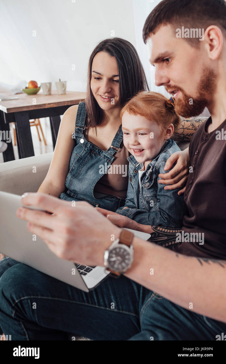 Smiling parents with daughter looking at laptop while sitting on sofa at home Stock Photo