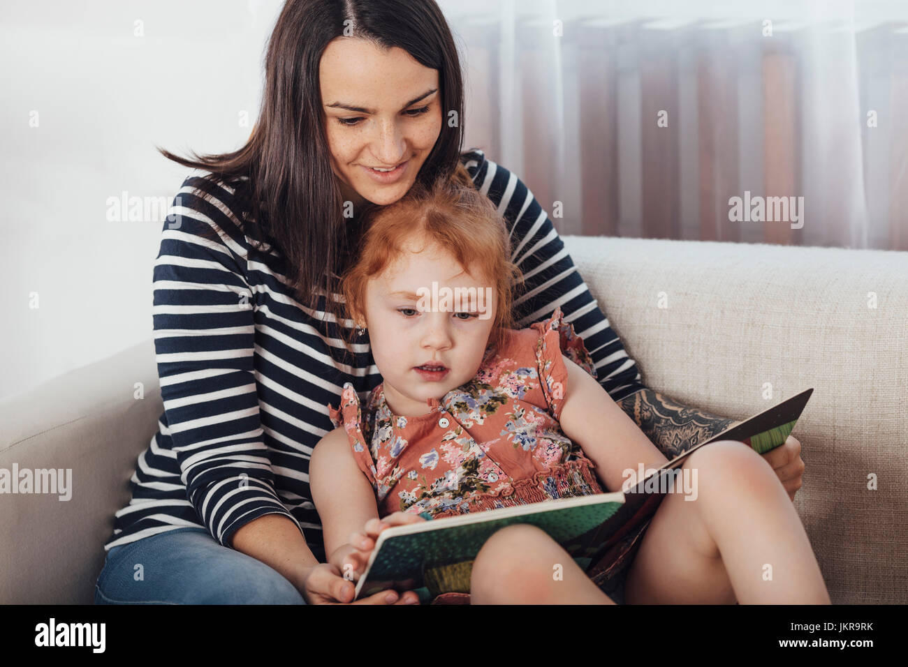 Smiling mother reading book to daughter while resting on sofa at home Stock Photo