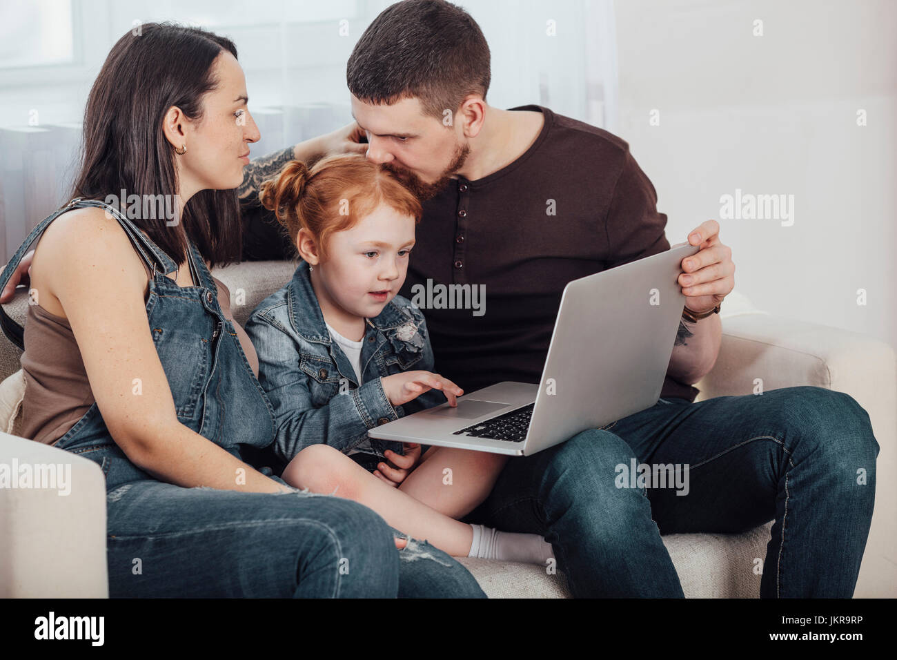 Woman looking at man kissing daughter while holding laptop in living room Stock Photo