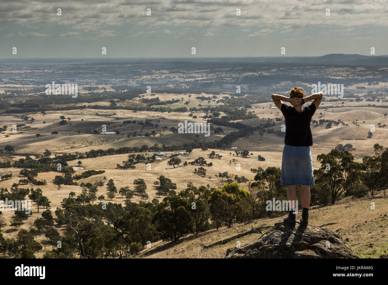 Rear view of woman standing with hands behind head on rock at mountain peak, Heathcote, Australia Stock Photo