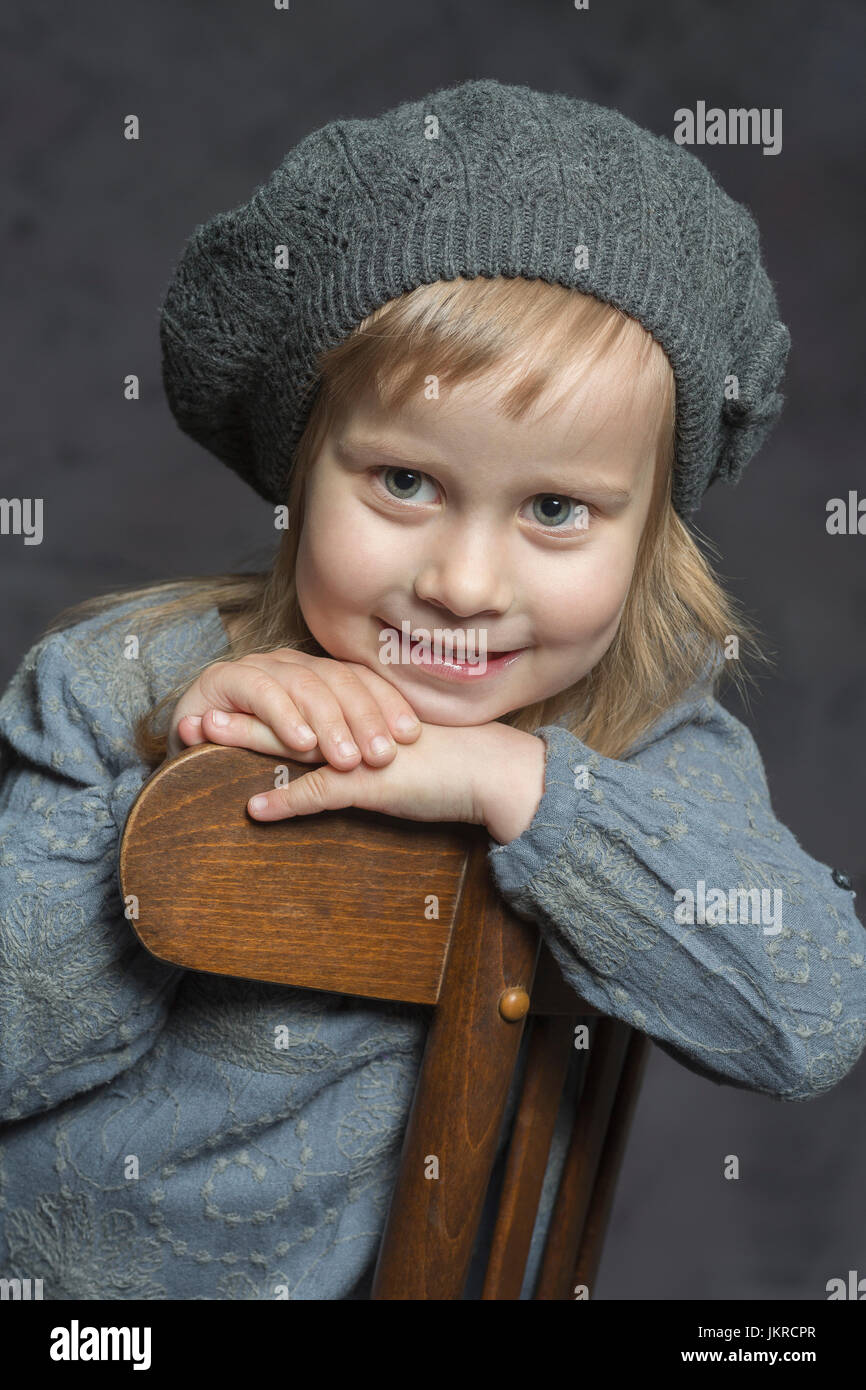 Cute smiling girl wearing knit hat sitting on wooden chair against gray background Stock Photo