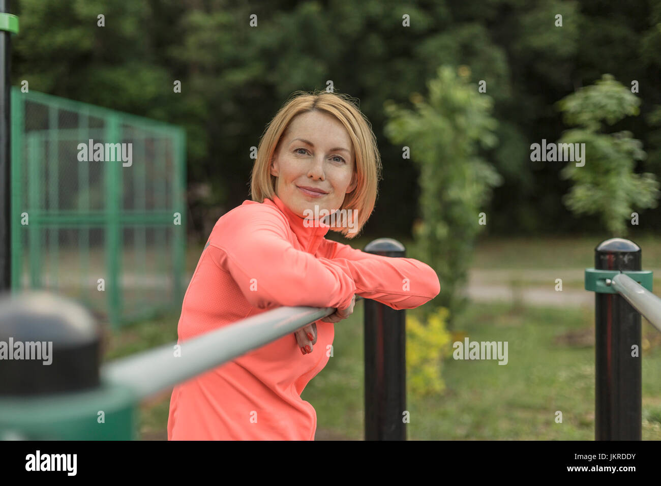 Portrait of smiling mature woman leaning on railing against trees at park Stock Photo