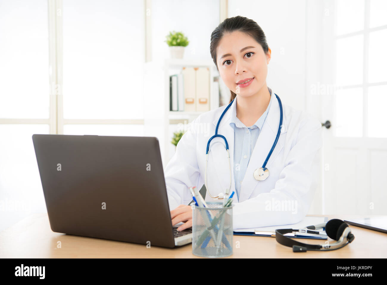 doctor woman sitting on her office with smile and looking to camera and mobile computer set on desk and medical equipment on her table typing with lap Stock Photo