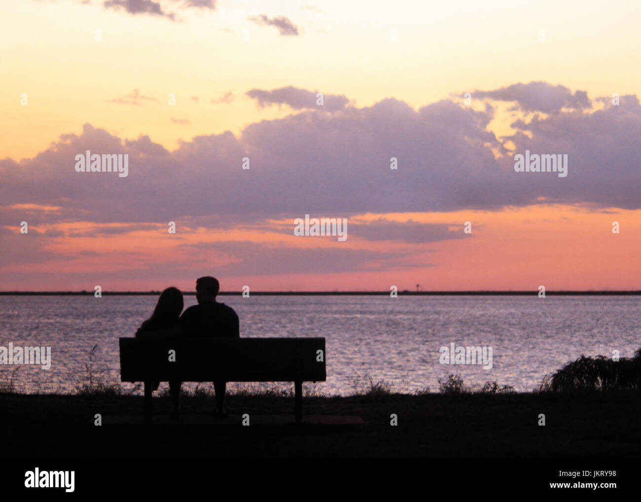 A couple sitting on a bench at a lake are silhouetted in the evening sunset. Stock Photo