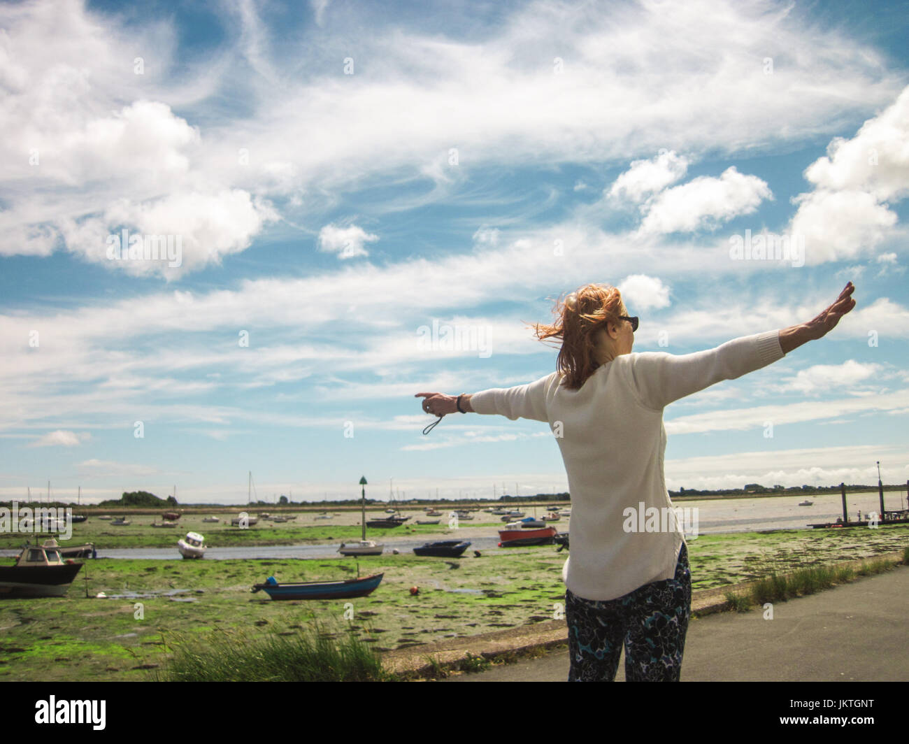 Rear shot of woman in white shirt gesturing with open arms at harbour view of small boats, on a cloudy summer's day in England. Space for text. Stock Photo