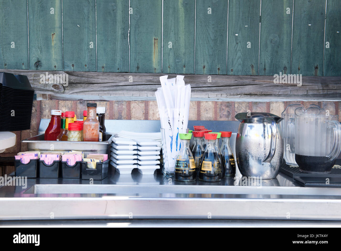 This is an image of a condiment station at an outdoor restaurant.  Condiments include, ketchup, hot sauce, pepper sauce, sweetner, straws, tea and wat Stock Photo