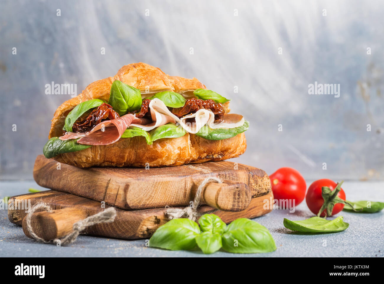 Croissant sandwich with smoked meat Prosciutto di Parma, sun dried tomatoes, fresh spinach and basil on stone textured grey background Stock Photo