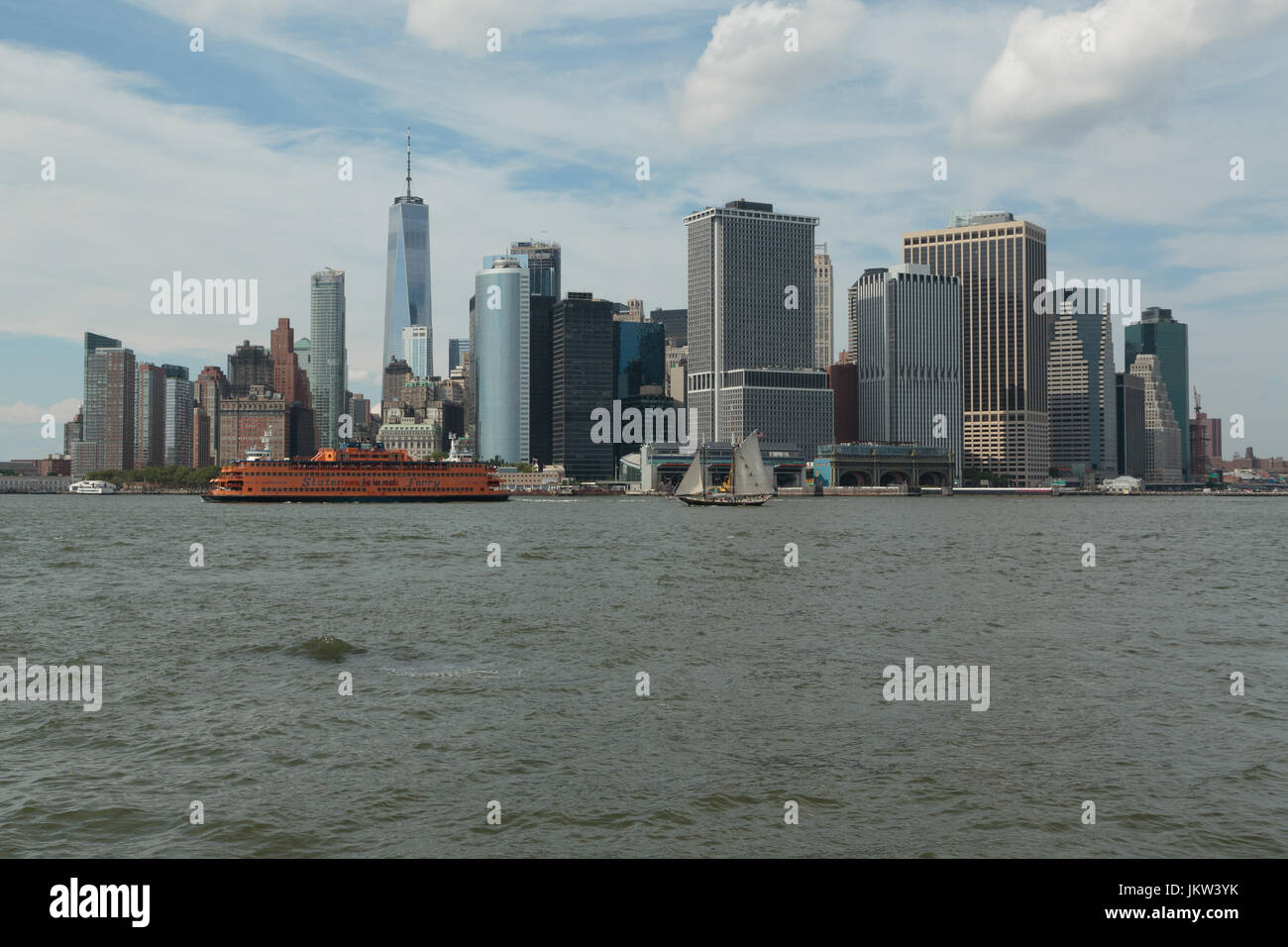 A photograph of Manhattan skyline, as seen from Governor's Island, New York City. The iconic orange Staten Island Ferry, and an old sailing ship can b Stock Photo